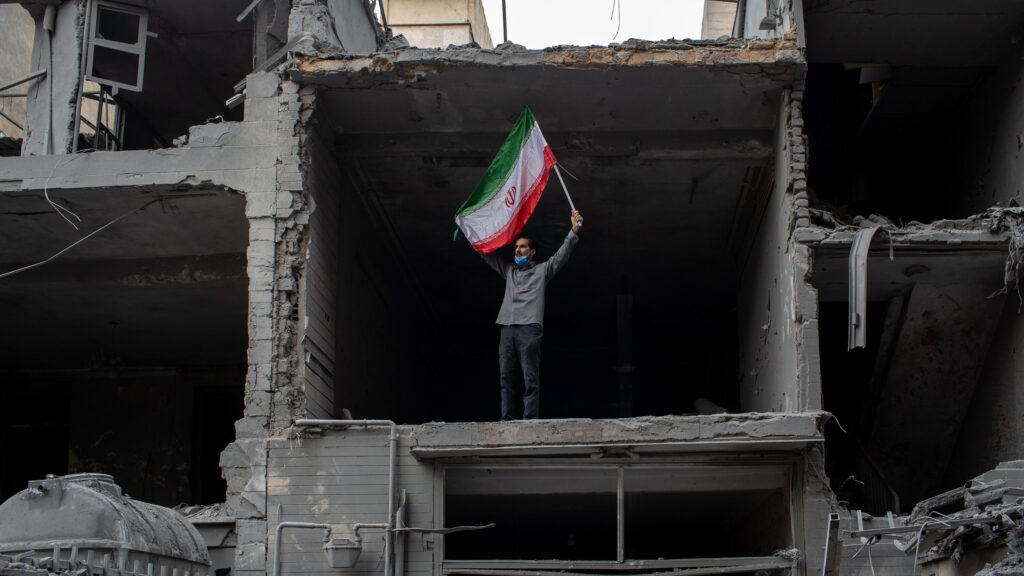 A man raises an Iranian flag in the ruins of a building in Tehran destroyed by U.S. and/or Israeli missiles. 