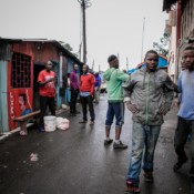 Several men stand outside a barbershop in a shack on a paved street.